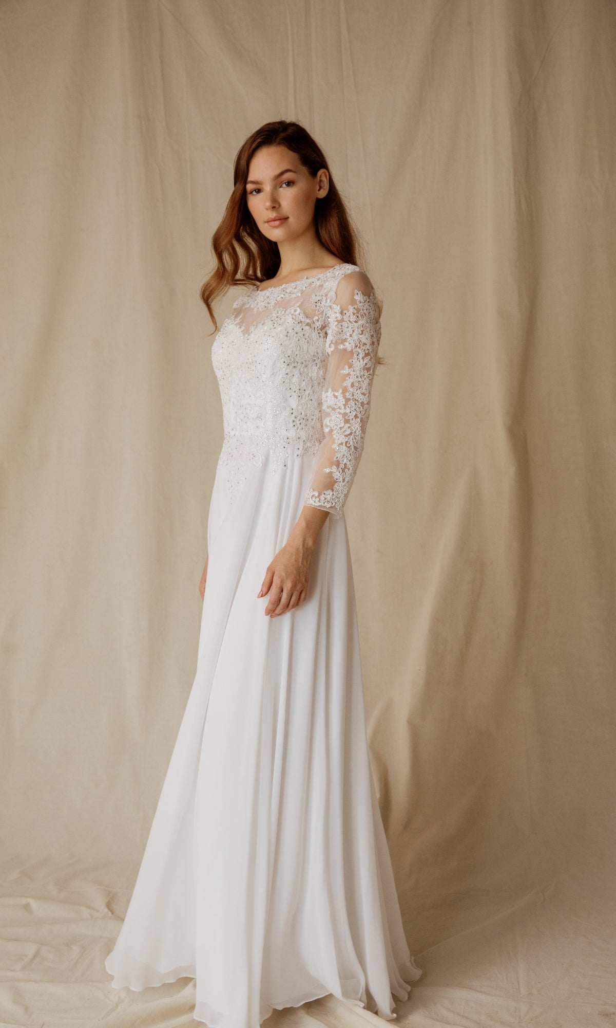 A woman models the Eureka 6909, a modest long-sleeve formal dress with lace accents, in front of a beige fabric backdrop—an elegant white gown ideal for special occasions.